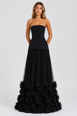 A woman modeling a strapless black formal gown with a structured bodice, flowing pleated skirt, and a dramatic ruffled floral hem against a light gray studio background.