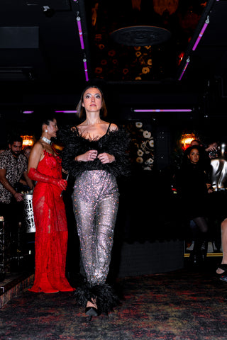 Model walking on a runway wearing a black top and silver pants with feathered shoes.