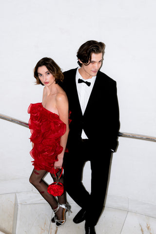 Man in a black tuxedo and woman in a red dress standing against a white wall.