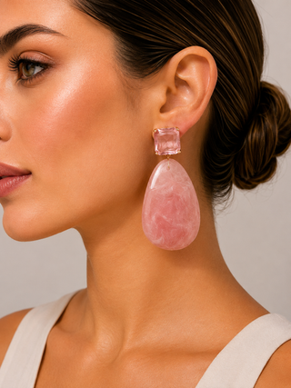 A close-up of a woman wearing a large pink teardrop dangle earring with a rectangular pink stud and gold-tone top, shown against a plain light gray background.