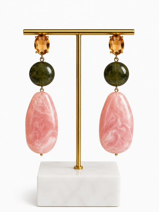 A pair of drop earrings displayed on a gold T-stand with a white marble base. Each earring features a round dark green stone, a teardrop-shaped pink marbled stone, and a small oval amber-colored stone at the top.