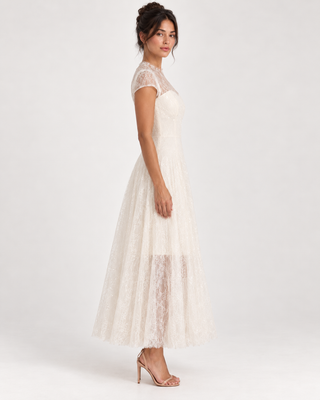 A woman modeling a sleeveless lace maxi dress in a warm ivory or cream tone, shown from the back three-quarter view against a clean light gray studio background.