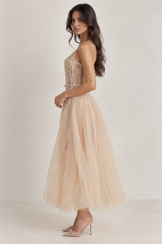 A woman modeling a sleeveless beige tulle maxi dress with a sheer lace bodice, photographed against a plain light beige studio background.