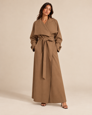 A woman modeling a long, wide-collar wrap coat in a warm taupe-brown color, styled with a matching tie belt and brown heeled sandals against a solid beige studio background.