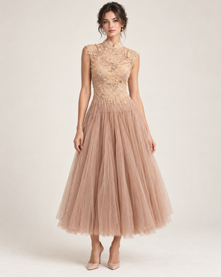 A woman models a sleeveless midi dress with a high mock neck, a fitted lace bodice, and a voluminous tulle skirt in a dusty blush pink color, paired with nude heels against a light beige studio background.