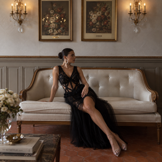 A woman wearing a black lace and tulle evening gown sits on a cream tufted sofa in a classic room with wall art, chandeliers, and terracotta tile flooring.