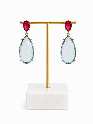 A pair of gold-tone drop earrings with marquise-cut deep pink gemstones at the top and large faceted light blue teardrop gemstones below, displayed on a gold T-bar stand with a white marble base against a white background.