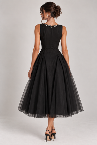 A woman modeling a sleeveless black midi dress with a pleated skirt and a beaded neckline, shown from the back against a light gray studio background.