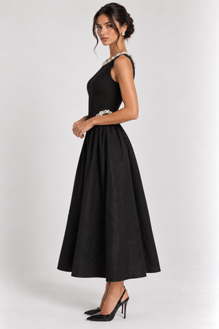 A woman modeling a sleeveless black midi dress with a structured bodice, embellished shoulder details, and a flared skirt, paired with black heels against a light gray studio background.