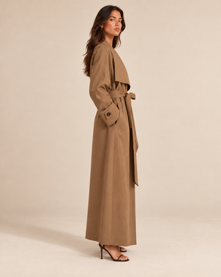 A woman modeling a long, wide collar belted trench coat in camel brown, shown from a side profile against a solid beige studio background.