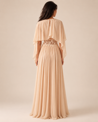 A woman shown from the back wearing a floor-length peach-beige gown with a draped cape-style back overlay, sheer embroidered waist detail, and a flowing pleated skirt against a warm beige studio background.