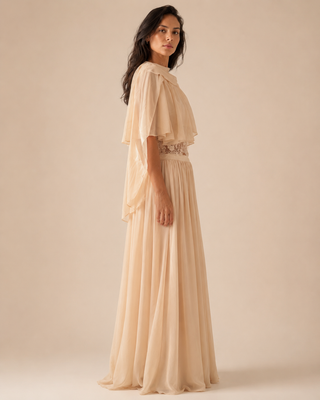 A woman modeling a floor-length cream or light beige chiffon gown with a draped cape-style sleeve, a fitted waist, and a pleated skirt, photographed against a plain warm beige studio background.