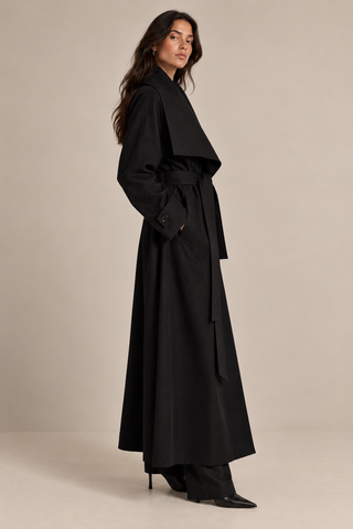 A woman modeling a long black wrap-style coat with a self-tie belt, shown in profile against a plain warm beige studio background.