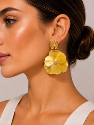 A close-up of a woman wearing a large, yellow flower-shaped drop earring with a textured gold top post, set against a neutral light gray background.