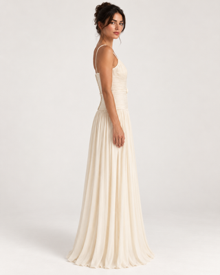 A woman modeling a floor-length cream or ivory sleeveless gown with thin straps, shown in side profile against a plain light gray studio background.