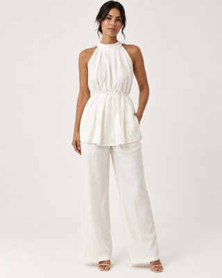 A woman modeling a white sleeveless halter top with a gathered peplum waist and matching wide-leg pants, photographed against a clean off-white studio background.