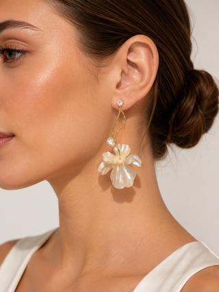A close-up of a woman wearing a dangling earring with a small clear stone stud and a large cream flower charm with layered pearl-like petals and a gold hook.