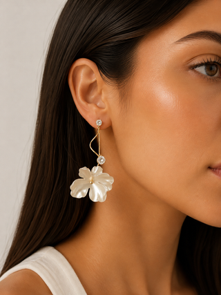 A close-up of a woman's ear wearing a dangling earring with a white flower-shaped drop and a curved gold wire detail, against a light neutral background.