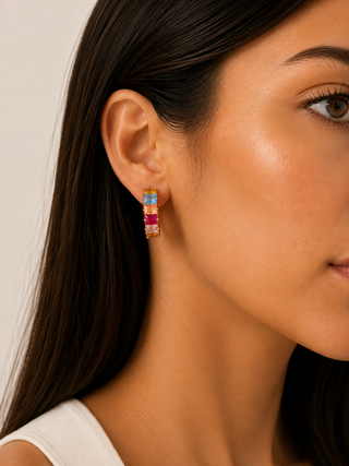 A close-up of a woman's ear wearing a small gold hoop earring with alternating blue, pink, and red gemstone-like segments.