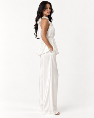 A woman modeling a white sleeveless jumpsuit with a peplum waist and wide-leg pants, shown from a side-back angle against a plain white studio background.