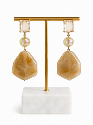 A pair of gold-tone drop earrings with clear rectangular crystals, iridescent round beads, and large organic-shaped tan stone pendants, displayed on a gold T-stand with a white marble base against a white background.