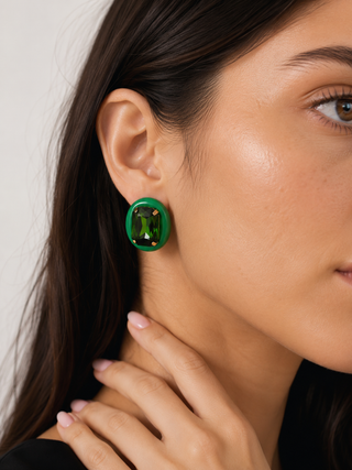 A close-up of a woman wearing a green rectangular stud earring with a cushion-cut green center and small gold corner accents, against a light neutral background.