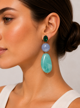 A close-up of a woman wearing a long drop earring with three stacked oval stones: a dark green top stone, a light periwinkle blue middle stone, and a larger marbled aqua-green bottom stone.