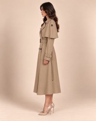 A woman modeling a mid-length beige trench coat with a belted waist and shoulder epaulettes, paired with clear high-heeled pumps, against a solid beige studio background.