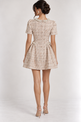 A woman viewed from the back wearing a short-sleeve tweed mini dress with a pleated skirt and pearl embellishments at the neckline, paired with beige high-heeled sandals against a light gray studio background.