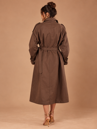 A woman viewed from the back wearing a brown trench coat with a matching waist belt, paired with gold high-heeled sandals, against a solid warm tan studio backdrop.