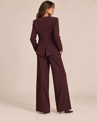 A woman wearing a dark burgundy two-piece suit with wide-leg trousers and a structured blazer, shown from the back against a solid beige studio background.