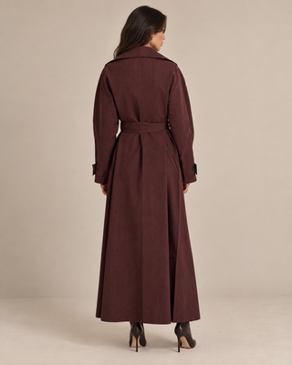 A woman viewed from the back wearing a long, belted trench coat in a deep burgundy-brown color, paired with dark pointed-toe heels, against a plain beige studio background.