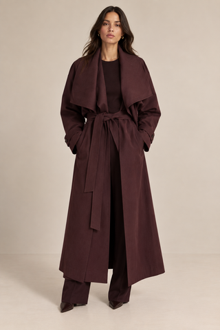 A woman stands against a plain beige studio background wearing a long dark plum-brown belted coat over a matching dark outfit.