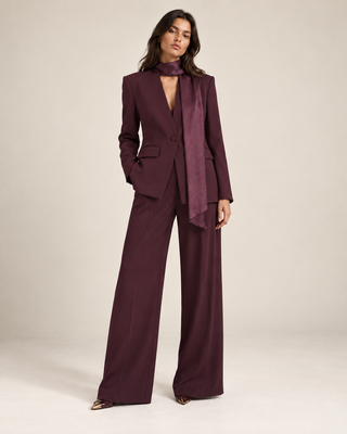 A model wearing a deep plum purple two-piece suit with wide-leg trousers, a matching satin neck scarf, and pointed-toe heels, standing against a neutral beige studio background.