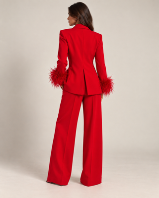 A model is shown from the back wearing a bright red tailored pantsuit with feathered cuffs on the blazer, set against a plain light beige studio background.