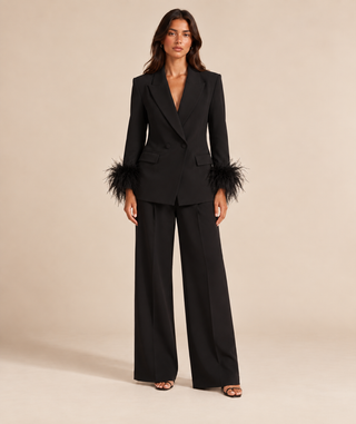 A model stands against a warm beige studio background wearing a black tailored blazer and matching wide-leg trousers. The blazer has notched lapels, a deep V-neckline, flap pockets, and dramatic feathered cuffs.