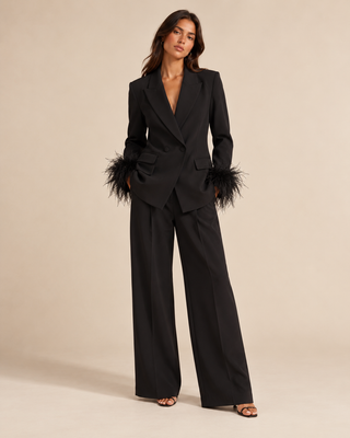 A woman models a black tailored blazer and wide-leg trousers set with dramatic feathered trim at the cuffs, photographed against a plain warm beige studio background.
