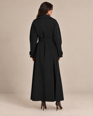 A woman viewed from the back wearing a long black trench coat with a matching waist belt, paired with dark pointed-toe heels, against a plain beige studio background.