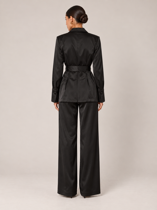 A model viewed from the back wearing a black satin two-piece suit consisting of a belted jacket and wide-leg trousers, set against a plain light gray studio background.