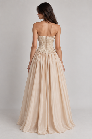 A woman shown from the back wearing a strapless formal gown with a structured, embellished corset-style bodice and a flowing chiffon-like skirt in a light champagne beige color, set against a plain light gray studio background.