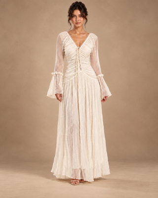 A woman modeling a long-sleeved cream maxi dress with a lace-up front and sheer flutter sleeves, standing against a solid beige studio backdrop.