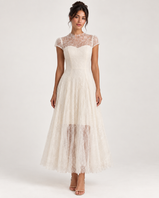 A woman modeling a cream lace midi dress with a sheer lace high-neck bodice, cap sleeves, and a layered tulle skirt, photographed against a plain light gray studio background.