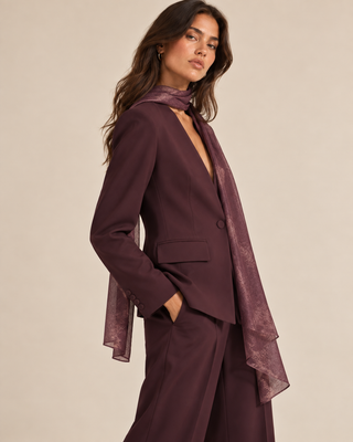 A woman models a deep plum purple tailored suit, featuring a structured blazer with a notched lapel, a matching skirt, and a sheer matching scarf draped over her shoulder, set against a solid beige studio background.