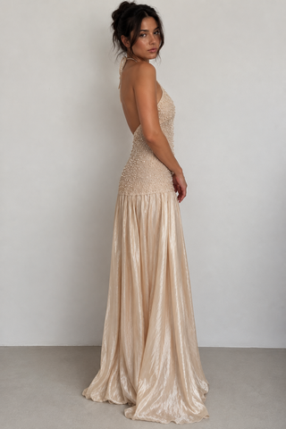 A woman modeling a champagne-colored, strapless maxi dress with a beaded bodice and a flowing satin skirt, shown from a three-quarters back angle against a neutral studio background.