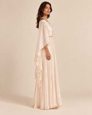 A woman modeling a floor-length cream or ivory maxi dress with draped cap sleeves, a pleated skirt, and a gathered waist, photographed against a plain warm beige studio background.