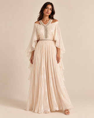 A woman modeling a floor-length off-the-shoulder maxi dress in a shimmering champagne beige with silver embellished neckline details and a matching beaded waistband, set against a solid warm beige studio background.