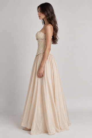 A woman modeling a strapless formal gown with a textured, beaded champagne bodice and a flowing chiffon skirt in a light beige color, photographed against a neutral light gray studio background.