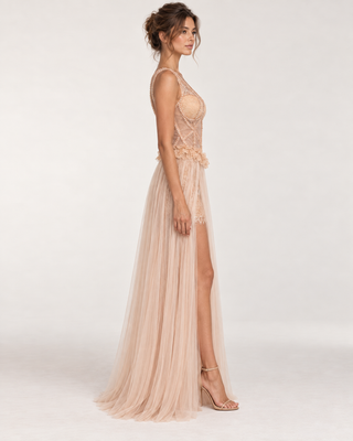 A woman modeling a floor-length nude-beige evening gown with a sheer lace bodice, floral applique at the waist, and a flowing tulle skirt with a high leg slit, photographed against a clean light gray studio background.