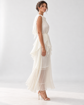 A woman modeling a sleeveless, tiered maxi dress in a soft ivory or off-white chiffon fabric, shown from a side-back angle against a clean, light gray studio background.