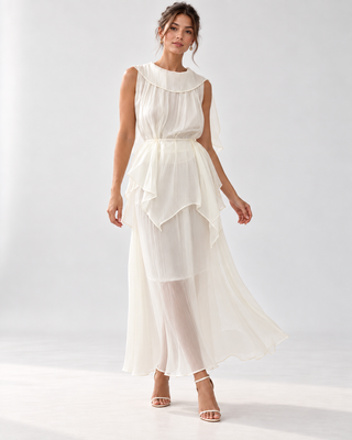 A woman modeling a sleeveless white midi dress with a layered, ruffled peplum-style skirt and a large ruffled collar, photographed against a clean white studio background.